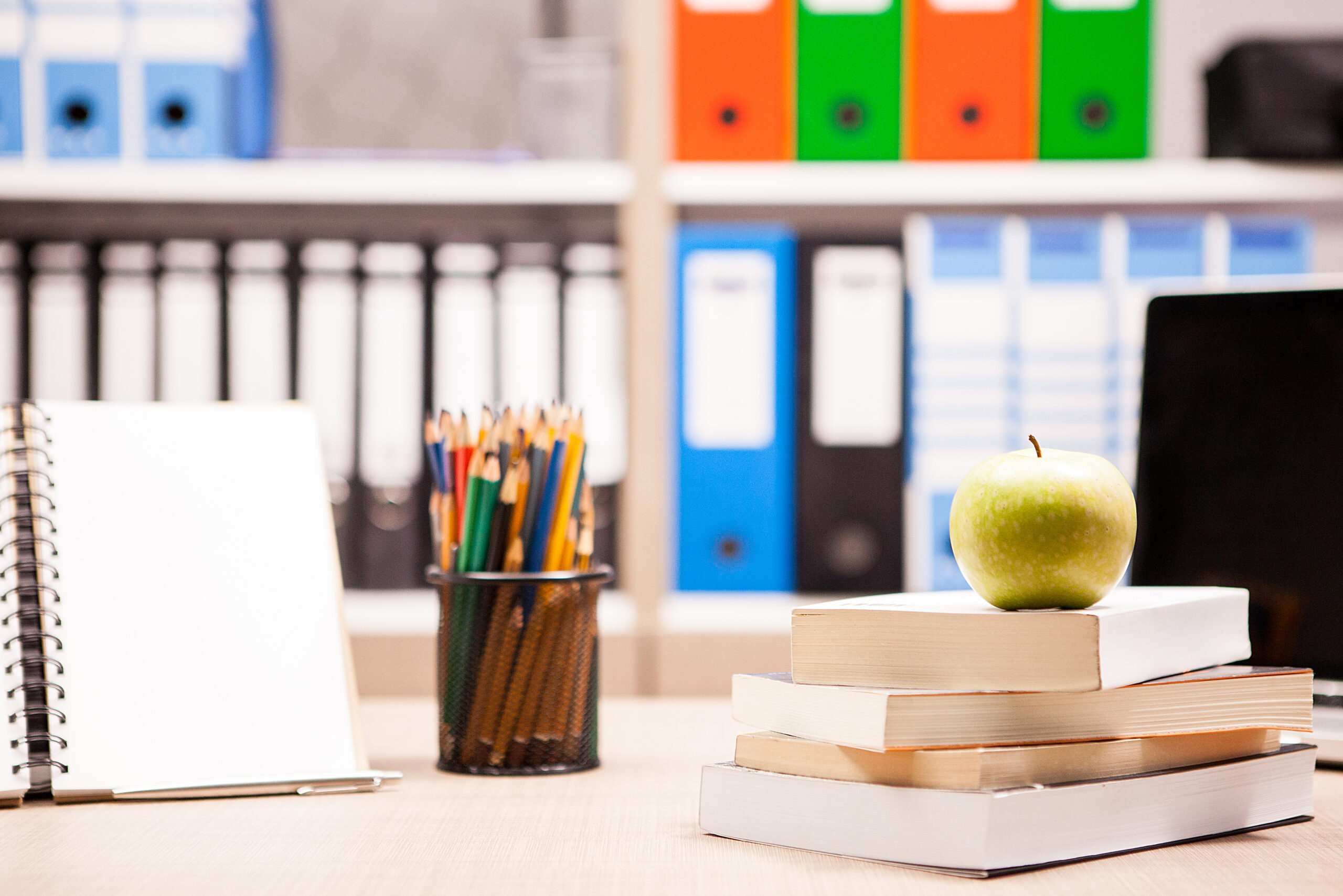 A green apple resting on a stack of books next to a spiral notebook and a cup of colored pencils, with colorful office binders suggesting the need to maintain teaching records blurred in the background.