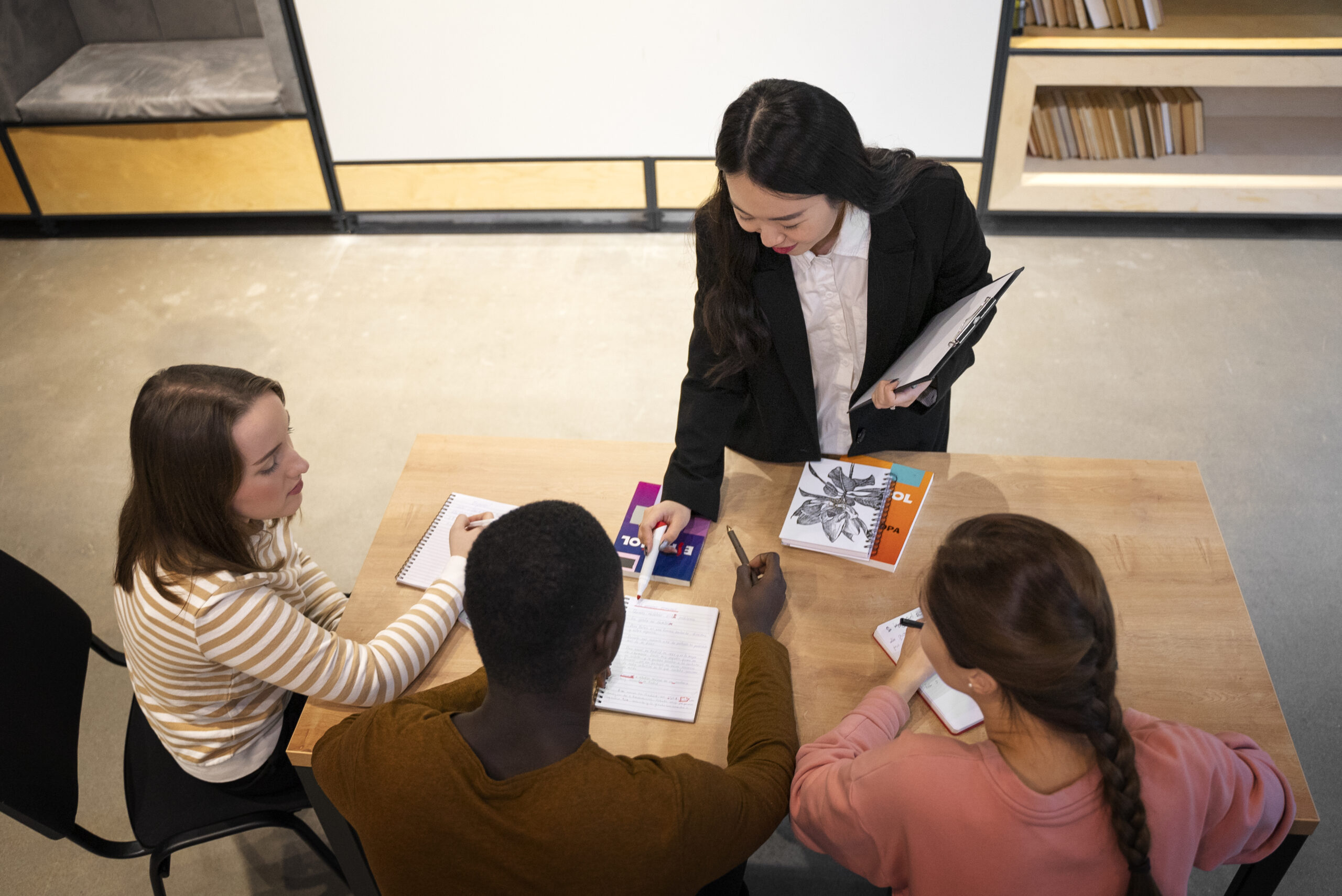 A high-angle shot of a diverse group of three students sitting at a wooden table while an Asian female teacher in a black blazer stands over them, helping to correct grammar or academic work. This depicts effective learning and engagement, supporting the keyphrase Eucto Campus teacher parent communication system.
