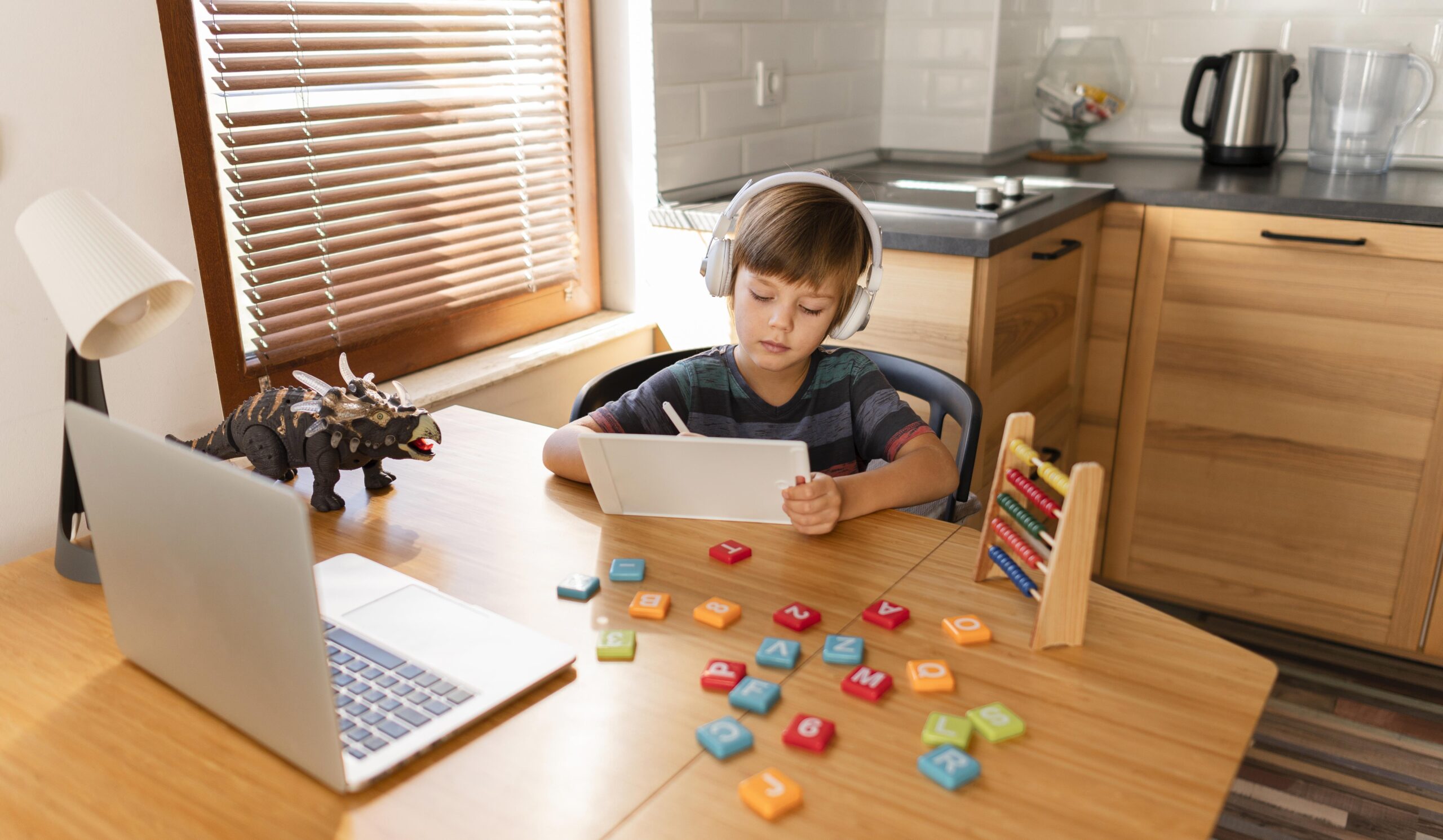 Focused young child wearing headphones and working on a tablet at a kitchen table, symbolizing parents' need to track child school activities during remote learning.