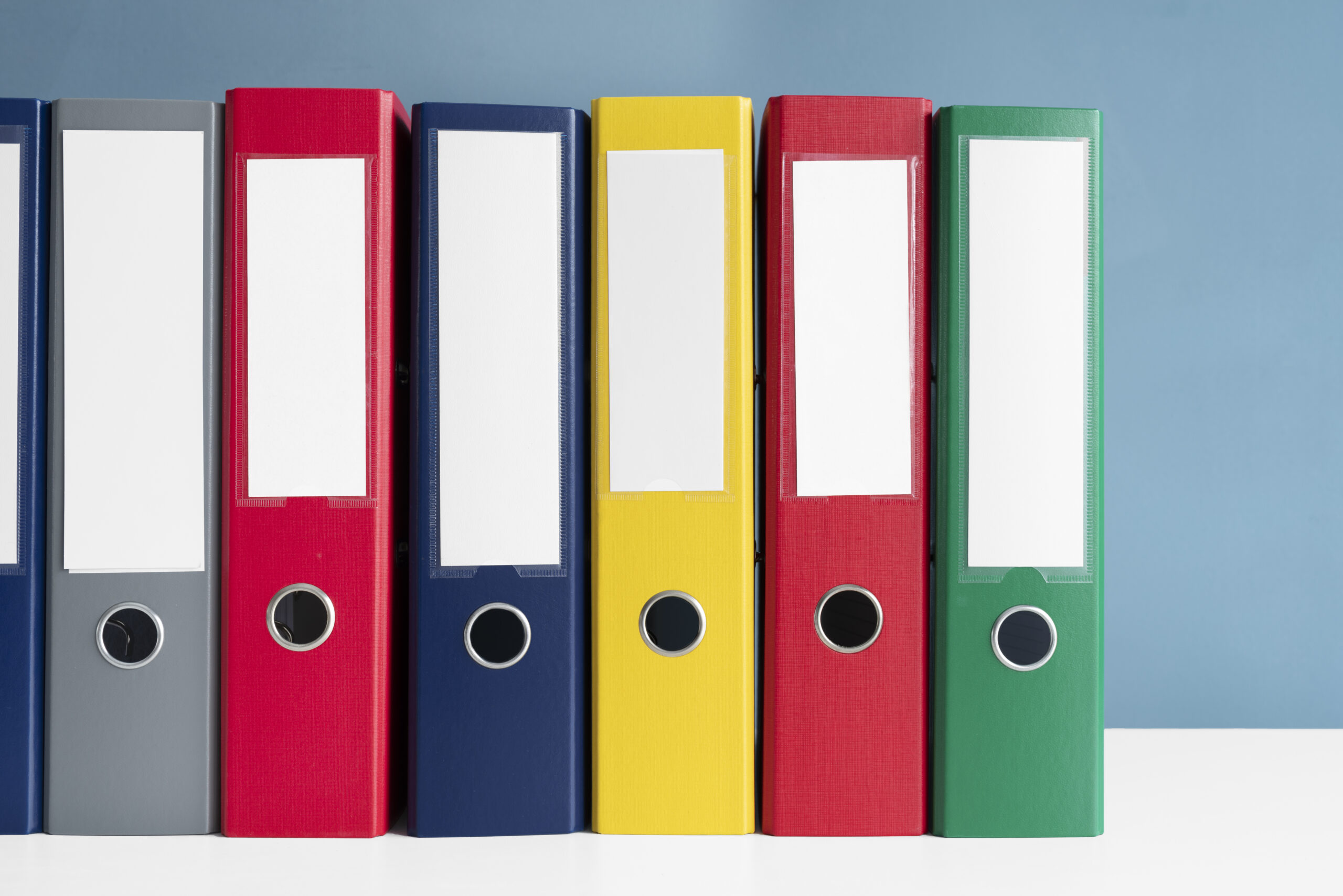 A neat row of colorful, labeled ring binders (grey, red, blue, yellow, red, green) used for storing and organizing documents.