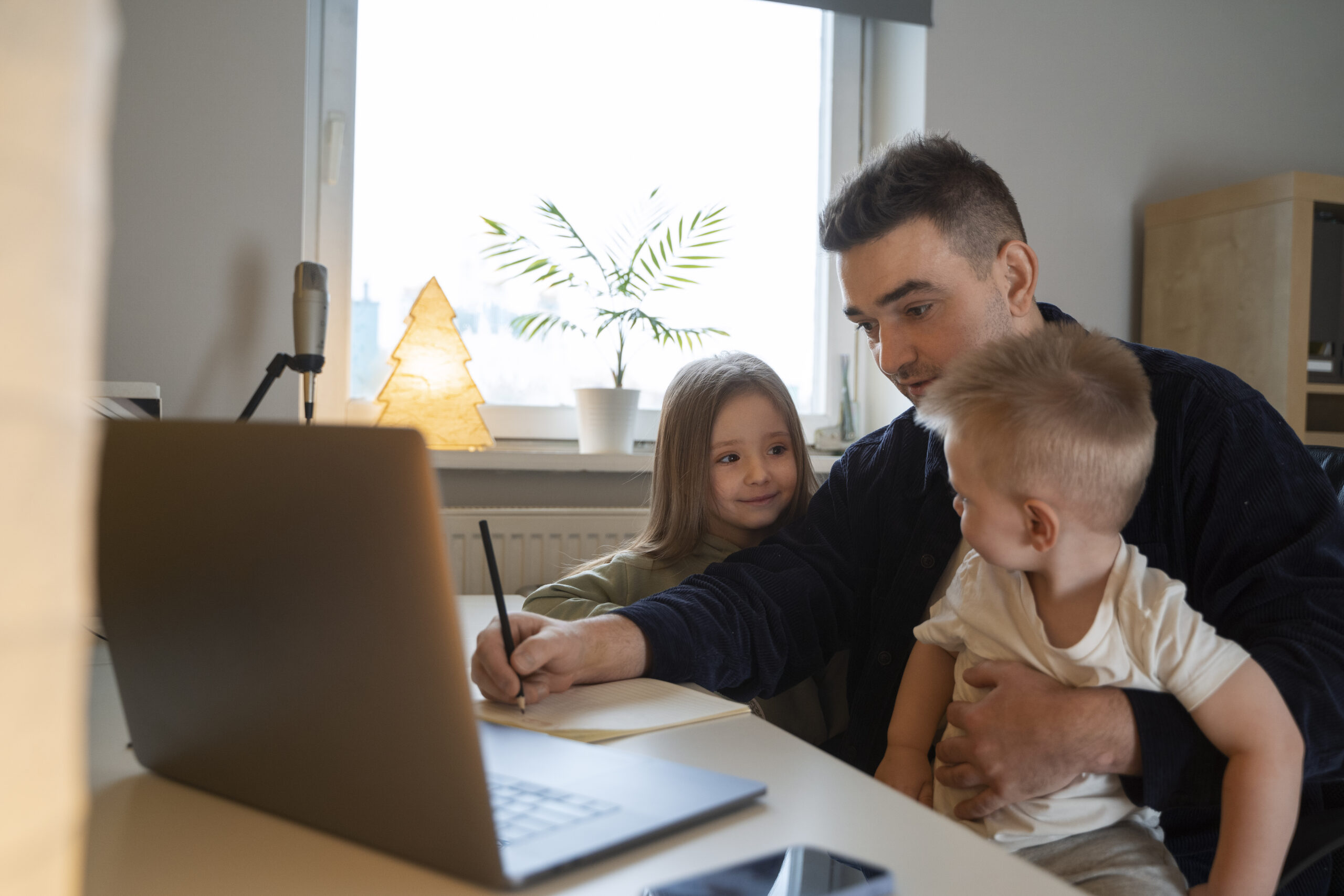 Father working on a laptop while holding a toddler and helping his daughter write, demonstrating ways to track child’s learning for working parents.