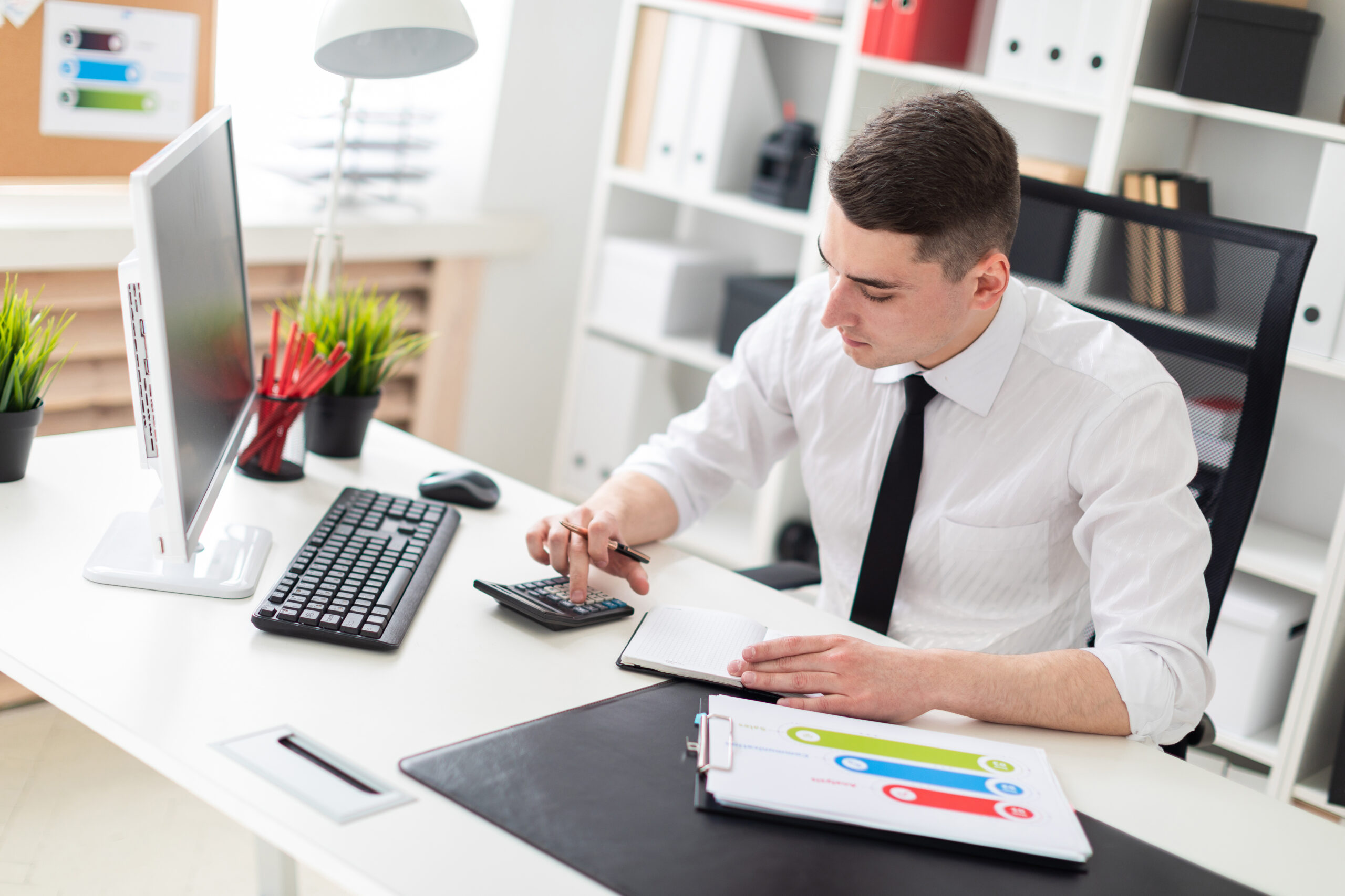 Focused young office worker calculating figures and reviewing financial documents at his computer desk, demonstrating methods to simplify school administration monitoring.